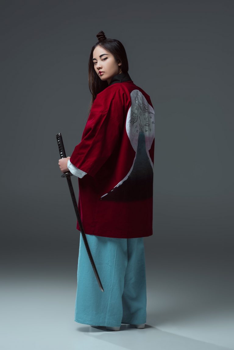 back view of woman in traditional japanese kimono holding katana and looking at camera on grey